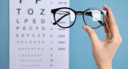 Woman holding glasses against eye chart on blue background, closeup. Ophthalmologist prescription